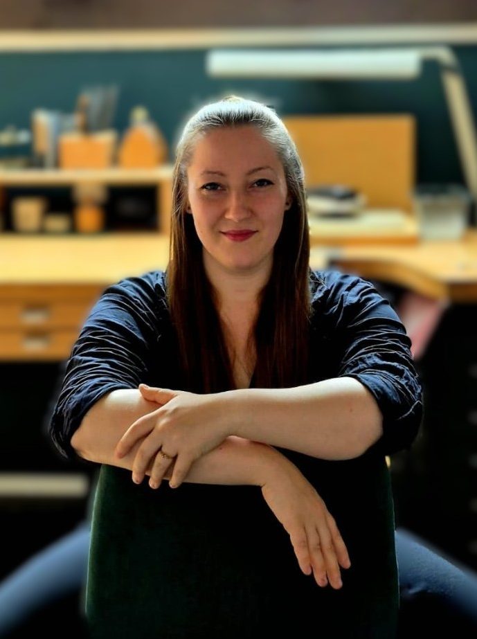 Inhaberin Juliane Golda sitzt lächelnd mit offenen langen Haaren in Ihrer Werkstatt vor Ihrem Werktisch.